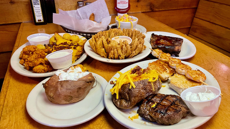 Multiple plates of food including steak, baked potato and Cactus Blossom on table at Texas Roadhouse rolls