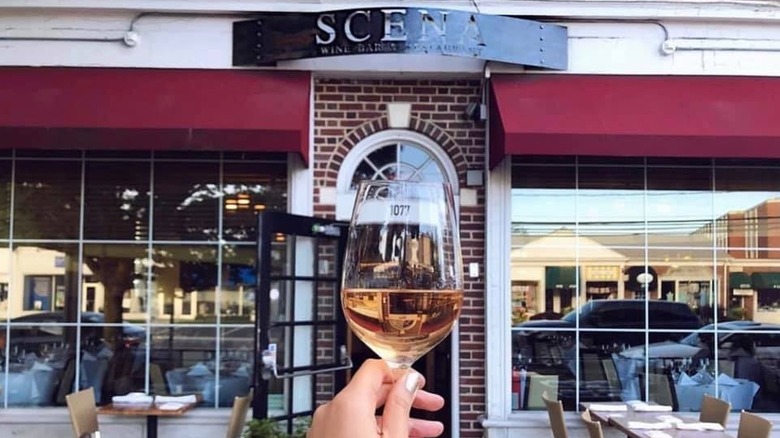 A hand holding a glass of white wine in front of the Scena wine bar patio and entrance