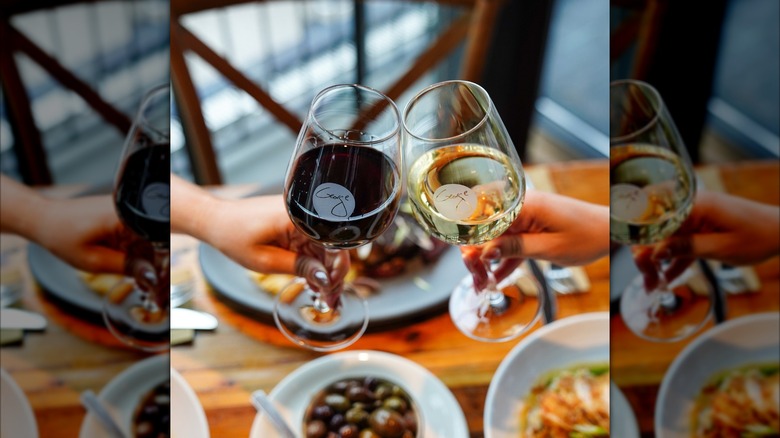 2 hands holding 2 glasses of wine clinking against each other. One glass has red wine, and the other, white, and both have the George logo. The background is blurred and there is a table with 3 different dishes.
