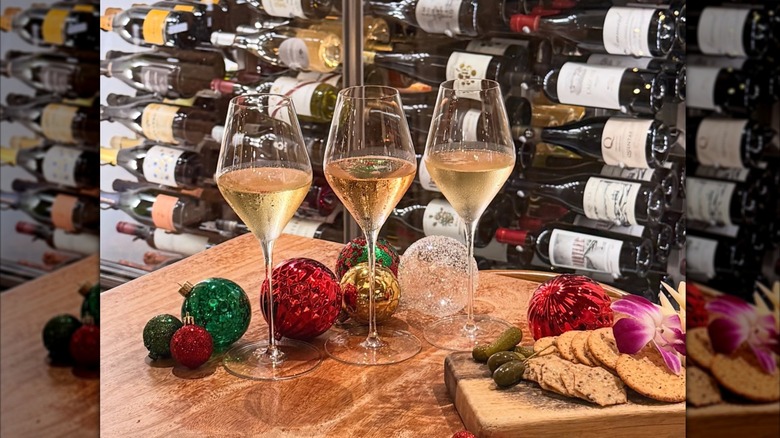 3 glasses of white wine on a table decorated with red, green, and gold christmas ornaments, and a charcuterie board. Wine rack with assorted wine bottles in the background.