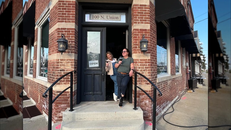 Two women stand in entryway of Westfield Wine vault in corner of brick building. They are laughing and holding glasses of red wine.