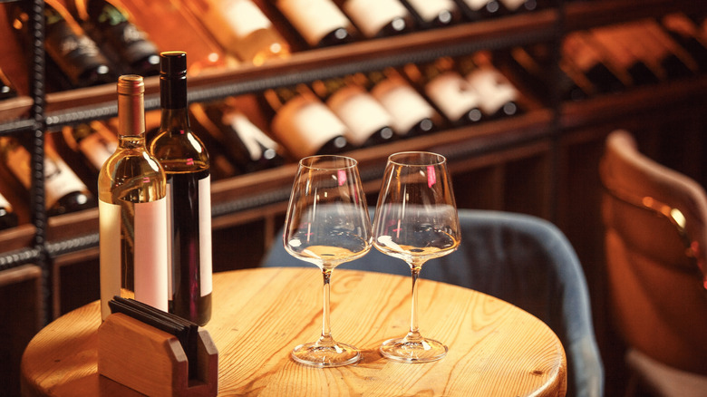 Two empty wine glasses on round, wooden table next to two bottles of red and white wine. Blurred background of more wine bottles on shelves.