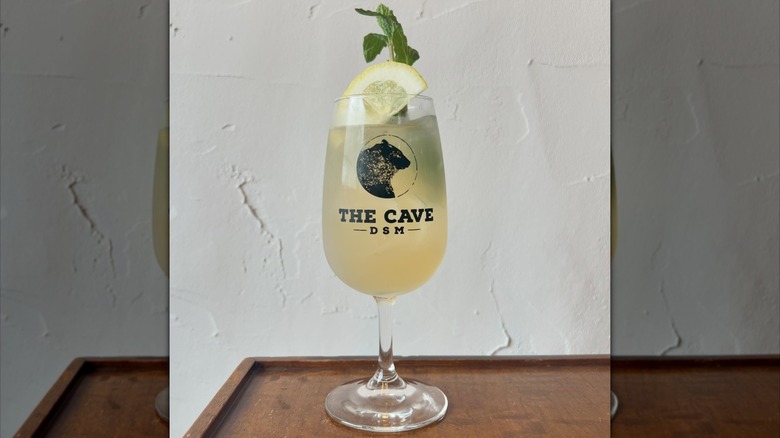 A glass of Rebujito Cocktail, with a lemon slice and mint leaves on top. The glass design has a bear, the logo of the wine bar, and the words "The Cave DSM." The cocktail is dry sherry with lemon soda.
