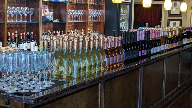 A counter at the bar full of sparkling wine and champagne bottles, with more glasses and bottles behind on shelves behind bartop