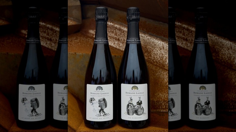 2 bottles of Domaine Lagille champagne that reads: vigneron a treslon depuis 1818. The bottle on the left has a label with a drawing of a man carrying a wooden barrel towards a tree. The one on the right has 2 humanoid rabbits sitting on a barrel each.