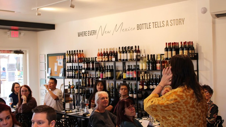 A group of people at NM Wine Studio. On one of the walls there are shelves full of wines and there is a phrase that reads: where every New Mexico bottle tells a story.