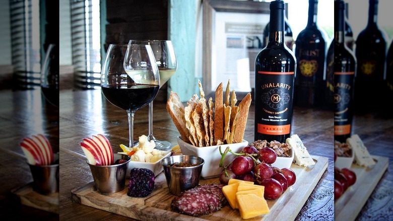 charcuterie board with snacks and grapes, alongside 2 glasses of wine and a bottle of Unalarity wine.