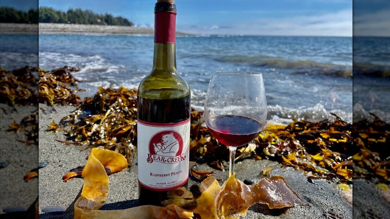 A bottle of Bear Creek Raspberry Peach wine next to a half-filled wine glass on a rocky beach with seaweed, waves, and a blue ocean in the background
