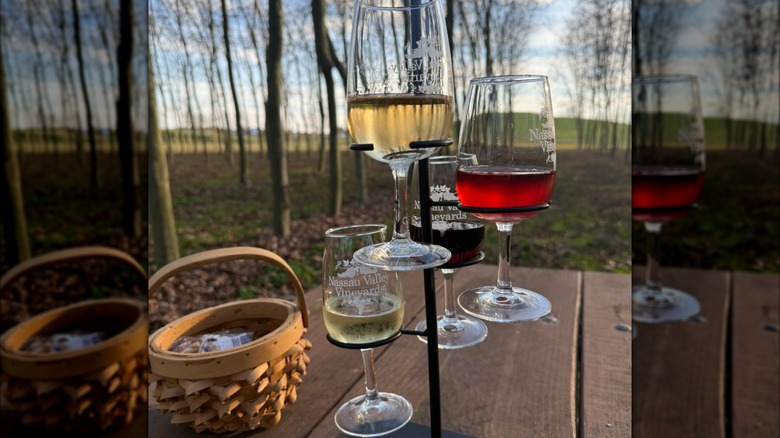 A wine flight on a black metal stand with glasses of white and red wine on a wooden picnic table overlooking a wooded setting