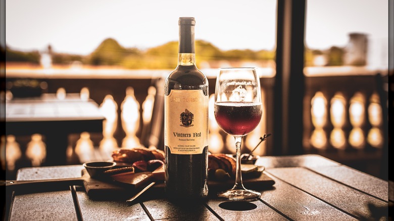 A bottle of red wine next to a filled wine glass on a wooden table with a charcuterie board on a balcony with a sunset view
