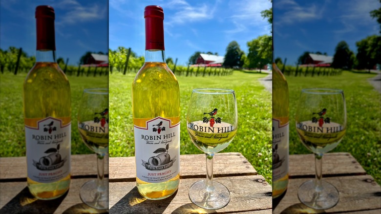 A bottle of Robin Hill Farm and Vineyards Just Peachy wine beside a wine glass on a wooden table, with a red barn and grape vines in the background