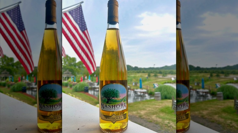 A bottle of Nashoba Valley Winery Vignoles white wine with an American flag and green hills in the background