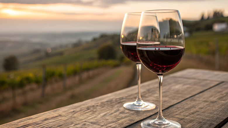 Two glasses of red wine on a wooden table overlooking hills of grape vines as the sun is setting