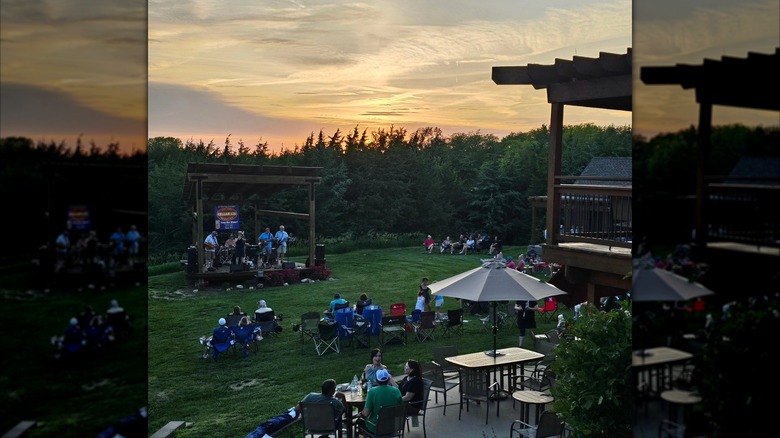 An outdoor sunset setting with people sitting on grass in lawn chairs with a band playing on a wooden stage and a patio overhang with tables and chairs