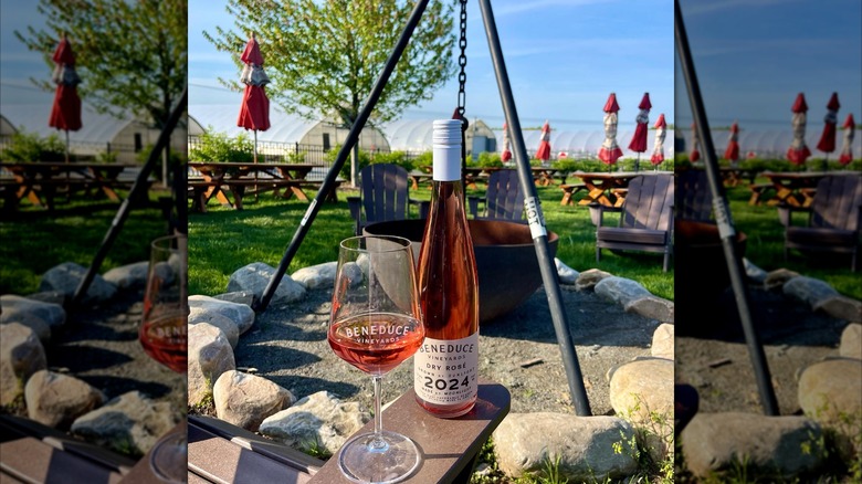 A bottle of Beneduce Vineyards 2024 Dry Rosé next to a half-filled wine glass in front of a fire pit with picnic tables, red umbrellas, and greenhouses in the background