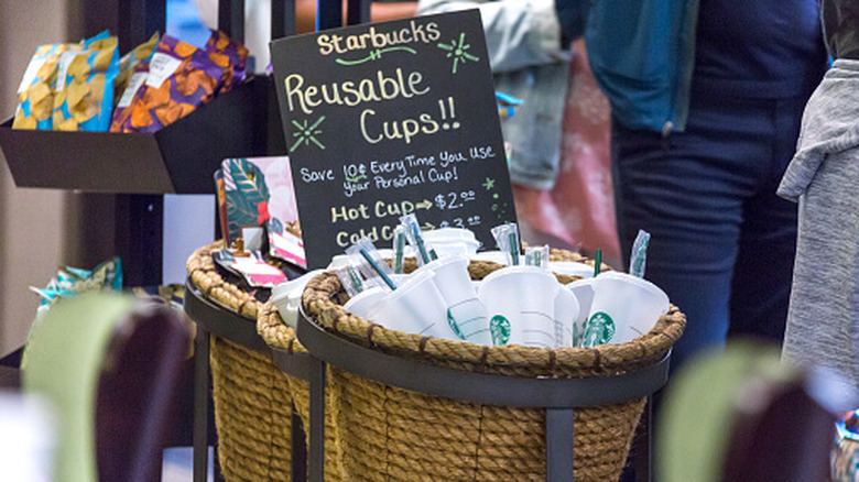 Basket with plastic reusable Starbucks cups next to customers waiting in line at Starbucks location.