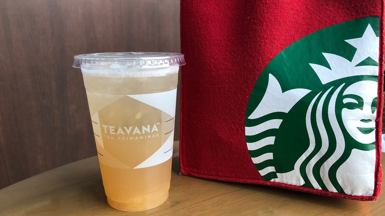 Plastic takeout Teavana cup with iced tea drink next to Starbucks bag on a wooden table.