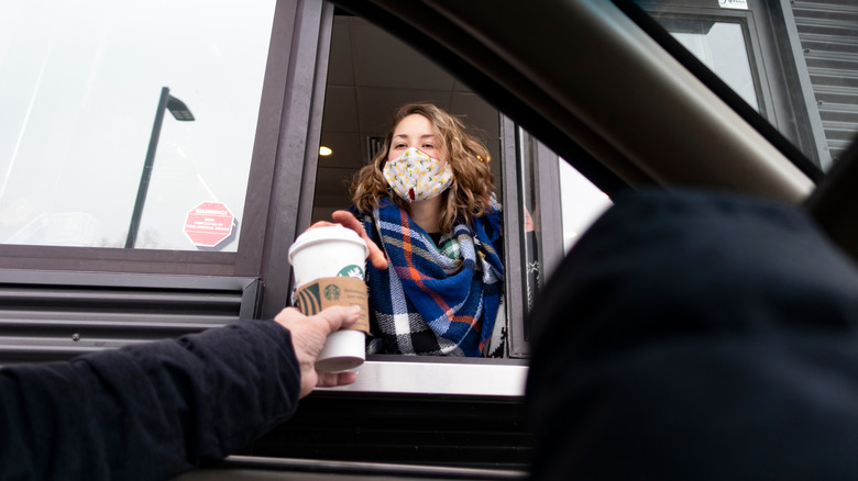 Masked Starbucks barista hands coffee through drive-thru window.