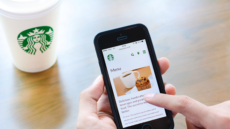 Hand holds a smartphone open to the Starbucks app menu page next to paper takeout Starbucks cup on table.