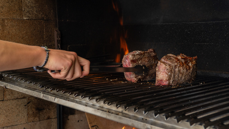 A hand using metal tongs to flip steak over on a grill