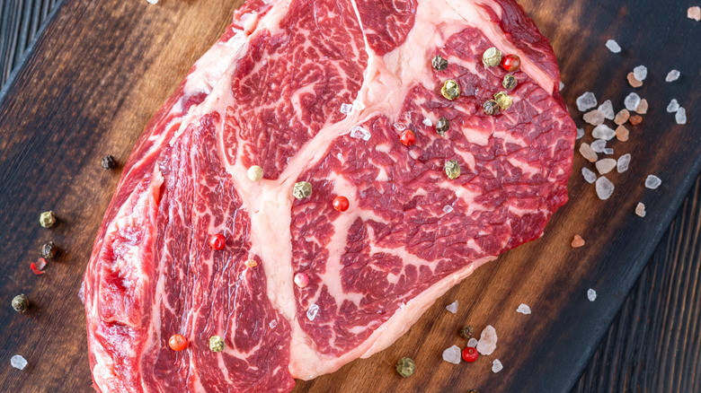 Raw ribeye on a wooden cutting board topping with coarse salt and whole peppercorns