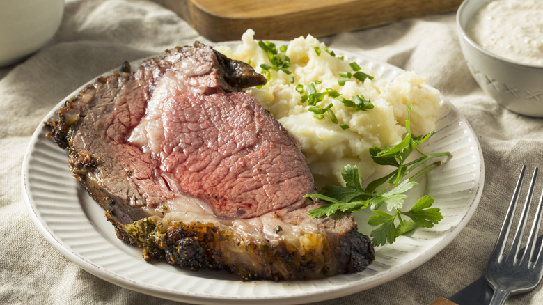Slice of prime rib on white plate with mashed potatoes and herbs.