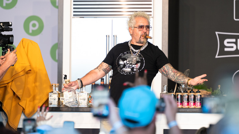 Guy Fieri faces audiences onstage with his arms outstretched, holding a clear bottle and a cigar between his lips