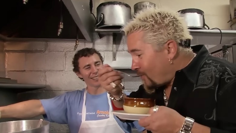 Guy Fieri smiling while eating a bowl of dumplings while the smiling chef looks on