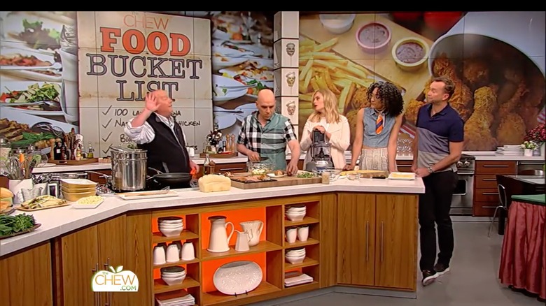 Mario Batali stands a little apart from his co-hosts Michael Symon, Daphne Oz, Carla Hall, and Clinton Kelly on the set of "The Chew"