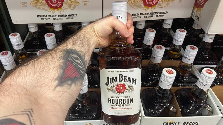 Hand holding bottle of Jim Beam bourbon in front of liquor display.