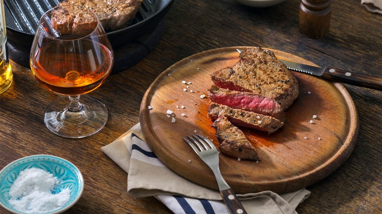 A sliced medium-rare steak on a wooden plate, next to a glass of bourbon.