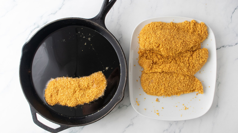 Breaded chicken cutlet frying in skillet next to more cutlets on plate