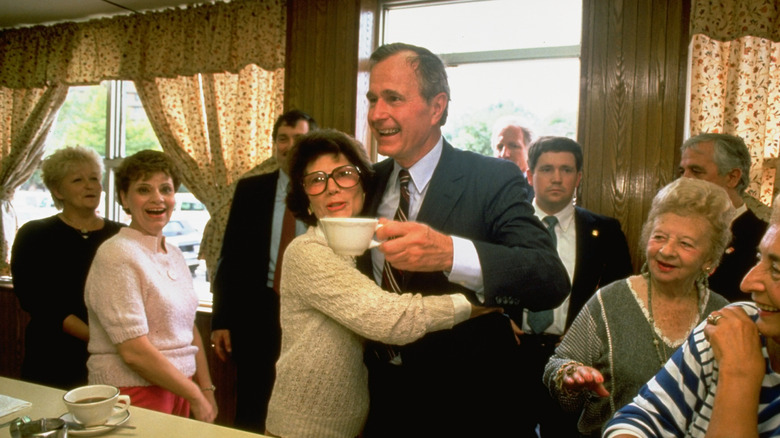 George H.W. Bush holding a cup of coffee and being hugged by a woman