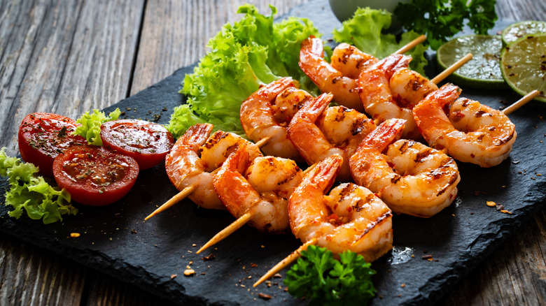 Shrimp on skewers with grill marks, served on a slate board, surrounded by lime, tomatoes, and lettuce.