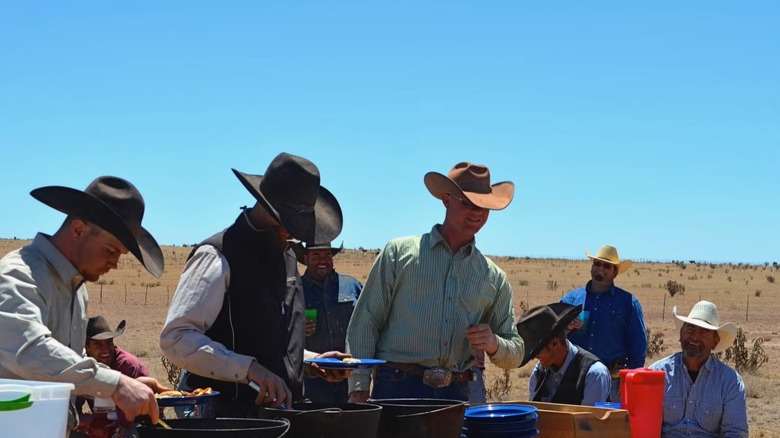 A group of cowboys gathered outdoors serving themselves plates of food from a table filled with cast iron pots
