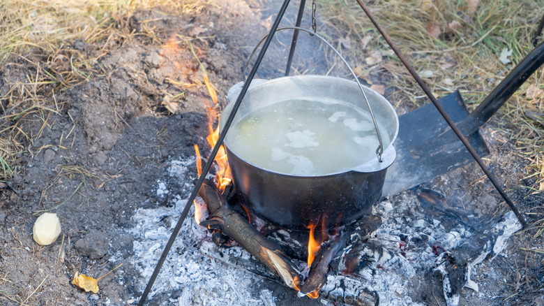 A shovel pushing burning firewood and ashy embers under a pot of water hanging from a metal camping tripod