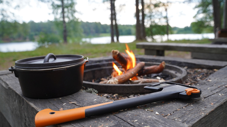 An Dutch oven outdoors near a lake on wooden surface with an axe and small campfire