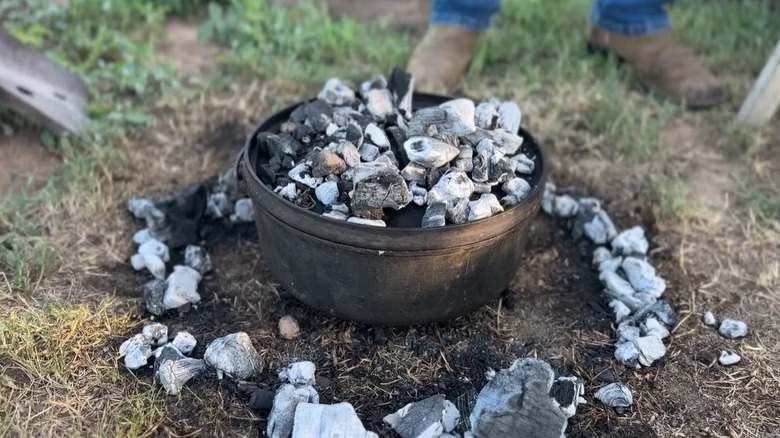 An outdoor cast iron Dutch oven on the ground with ashy coal around it and on top of its lid