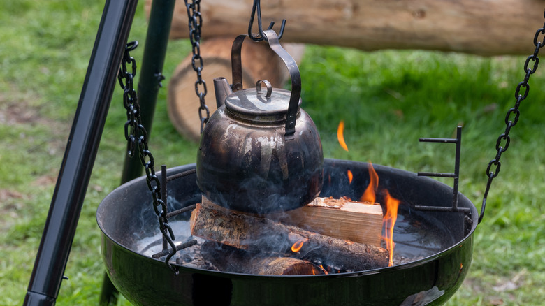 A metal coffee pot hanging from a tripod over a pan flaming firewood