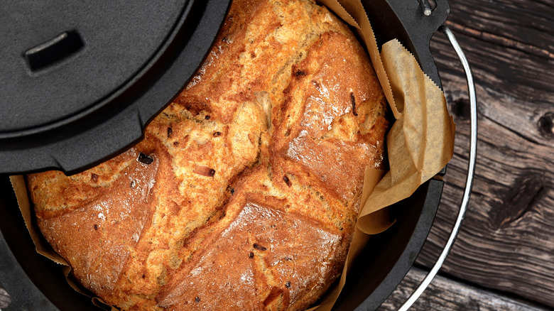 Baked bread inside in a paper-lined traditional cast iron Dutch oven with the lid partially closed on wooden surface