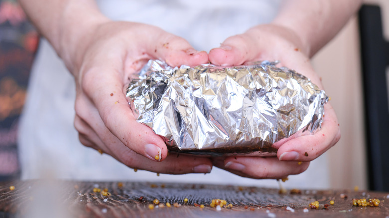Close up of person holding seasoned meat wrapped in foil over wooden surface