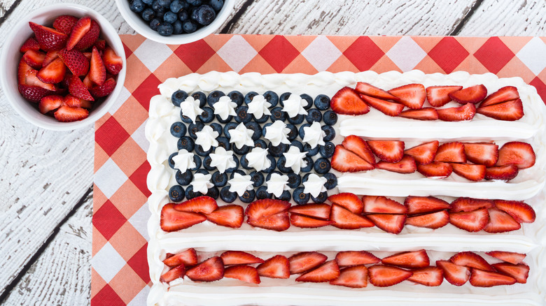 American flag cake topped with fruit on a plaid place mat with bowls and strawberries and blueberries