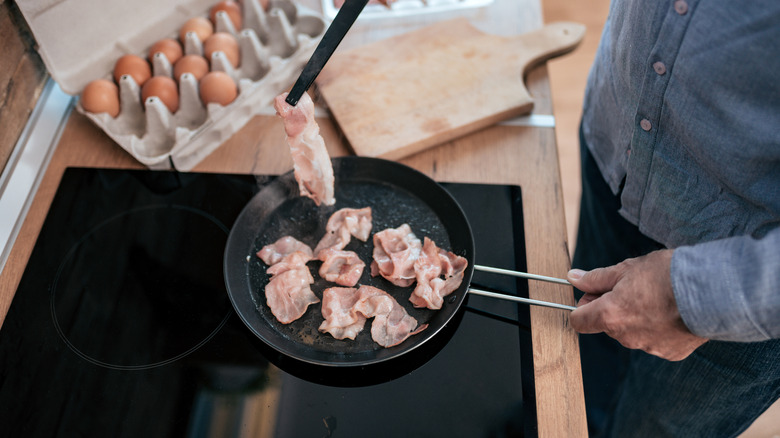 Person frying bacon on stovetop with carton of eggs nearby.