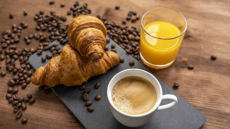 Coffee cup and two croissants on black slab, plus glass of orange juice and coffee beans on wood table.
