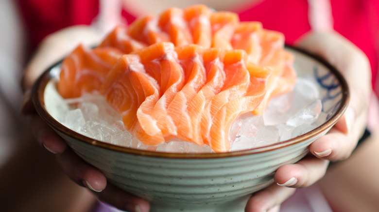 Hands holding bowl of sashimi layered over ice.