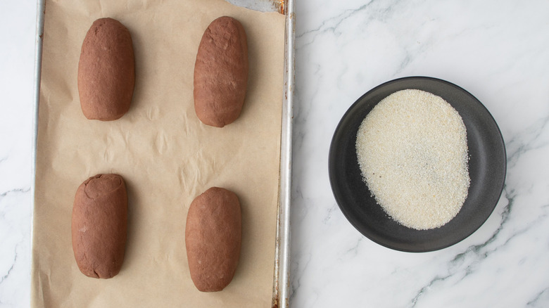 Brown bread loaves on baking sheet next to bowl of cornmeal