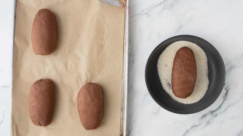 Brown bread loaves on baking sheet next to loaf in bowl of cornmeal