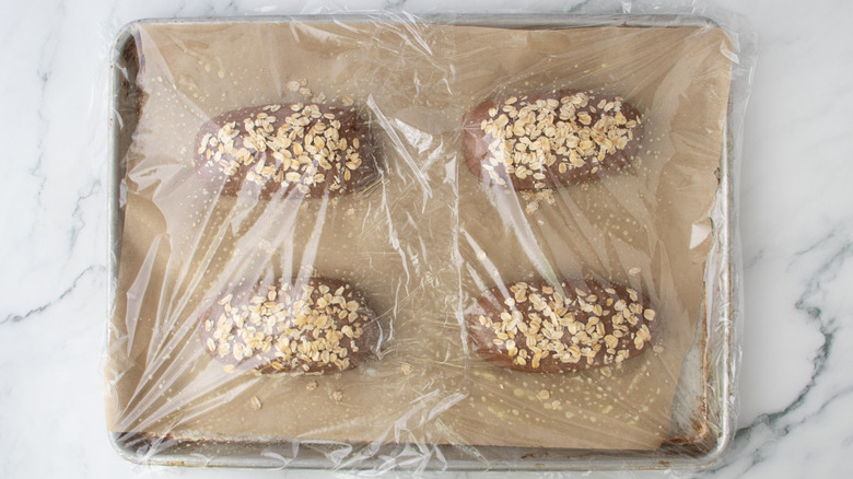 Brown bread loaves on baking sheet covered in plastic wrap