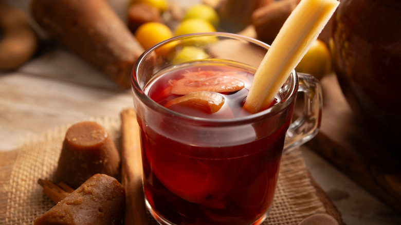 A glass of hot ponche nacideño with a stick of fresh sugarcane in it, surrounded by cinnamon sticks and piloncillo