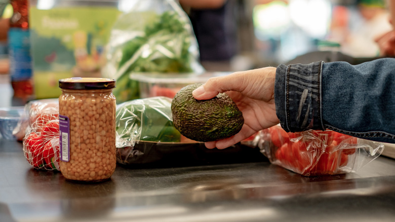 Hand placing avocado on grocery store conveyor belt with other food items.